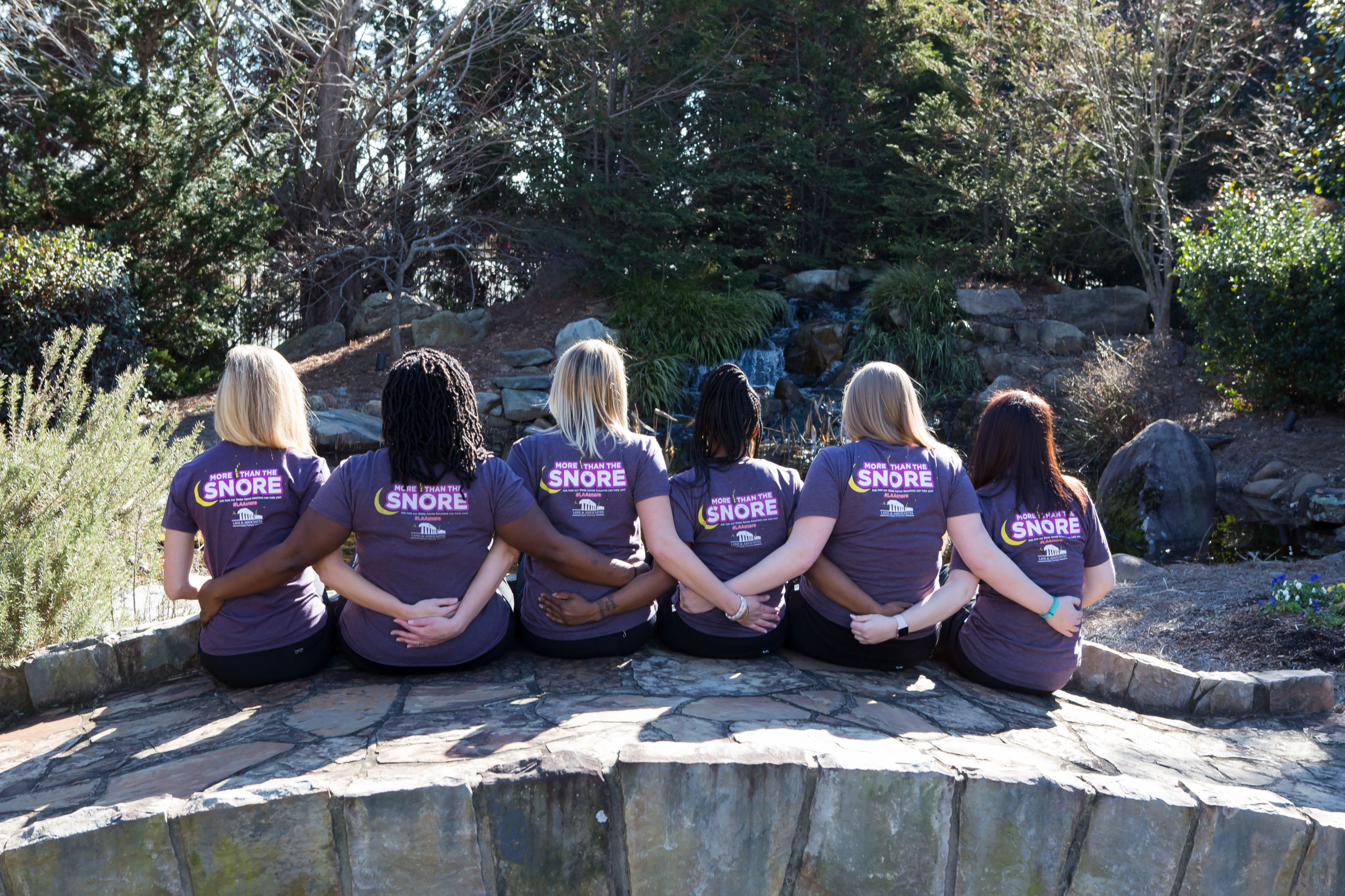group of women sitting on a bridge in Garner NC wearing More Than the Snore purple t-shirts backs toward camera
