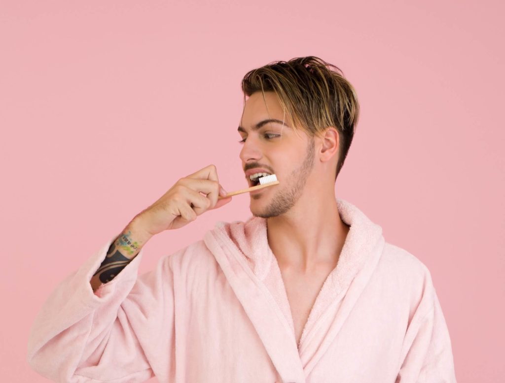 Adult hipster in pink bathrobe with pink walls brushing his teeth with tan toothbrush